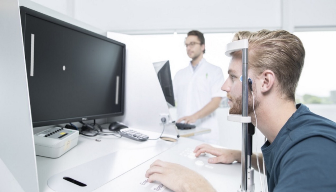 A participant with their head in a chin rest in front of a monitor, taking a NeuroCart test while a researcher watches